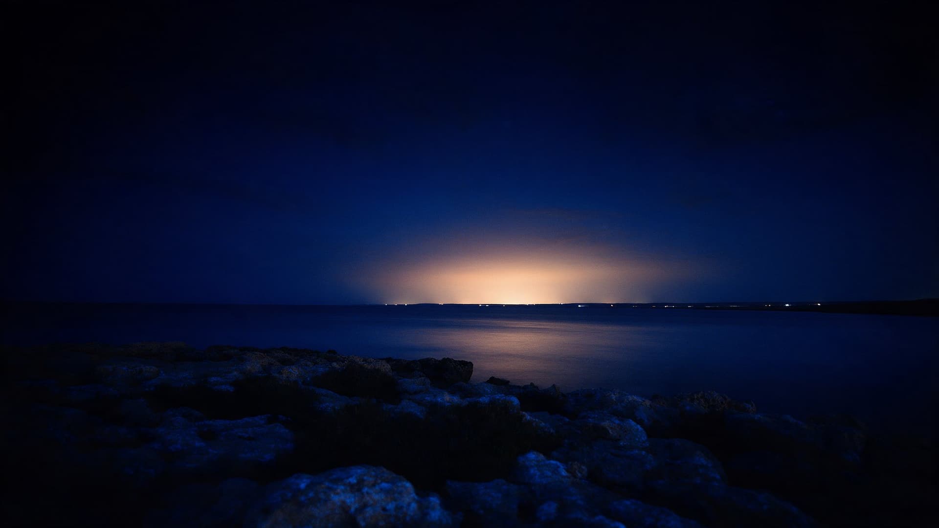 Felsige Meeresküste bei Nacht, am Horizont ein warmes Licht
